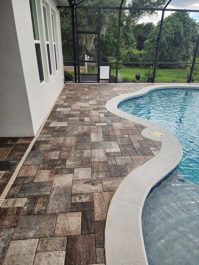 Backyard swimming pool with herringbone patterned pavers, white pool deck edge, screened enclosure, and green landscape views