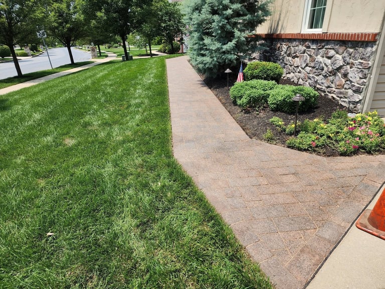A well-maintained residential property featuring a manicured green lawn, brick paver pathway, and landscaped flower beds with stones against a house foundation.