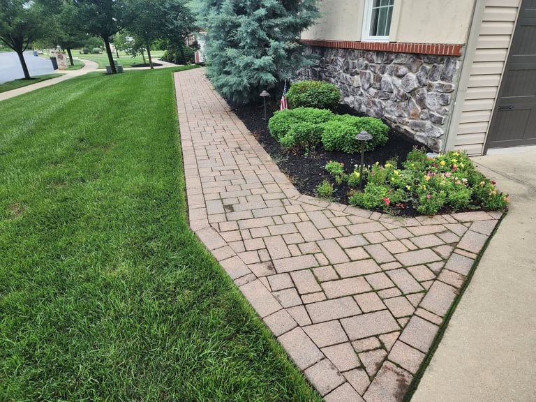 Residential walkway with brick pavers leading to house entrance, bordered by manicured lawn and landscaped shrubs